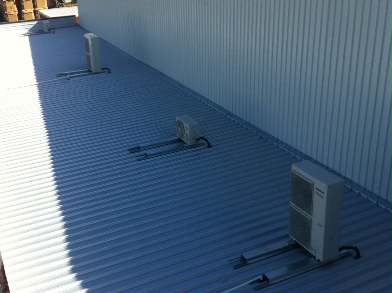 A row of Panasonic split system condensers mounted on steel rails along a commercial colorbond roof